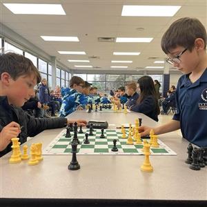 Two students focus on making their next move on the chess board. Others are in the background, playing in the same tournament.